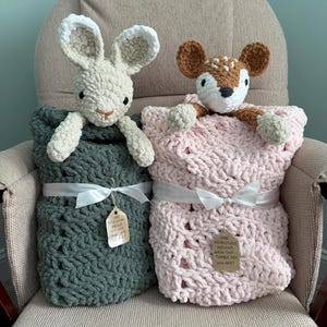 May include: Two crocheted plush toys, a cream-colored rabbit and a brown and white fawn, each tucked into a chunky knit blanket. The dark green blanket and light pink blanket are tied with white ribbons and feature tags with care instructions.