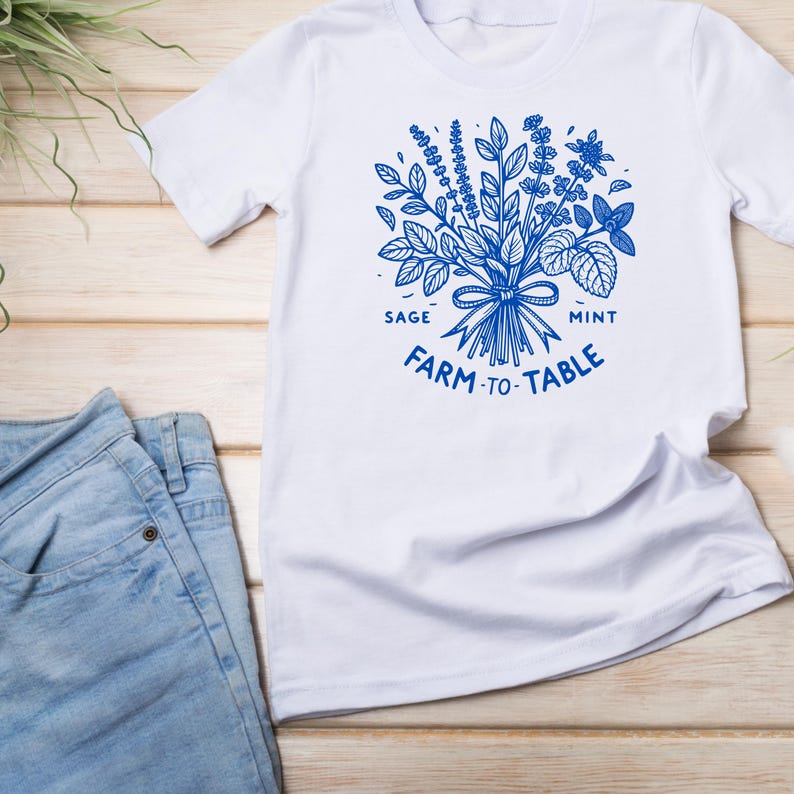 Może przedstawiać: Biała koszulka z niebieską ilustracją zi&oacute;ł, w tym szałwii i mięty, związaną kokardą. Tekst "FARM -TO-TABLE" jest wydrukowany poniżej. Jasnoniebieskie dżinsy są widoczne po lewej stronie.