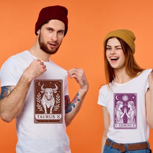May include: Two white t-shirts with zodiac signs printed on them. The shirt on the left has a brown bull with the text "TAURUS ♉" below it. The shirt on the right has a purple image of two women with the text "GEMINI ♊" below it.
