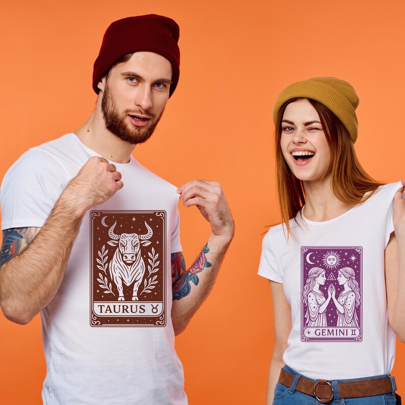 May include: Two white t-shirts with zodiac signs printed on them. The shirt on the left has a brown bull with the text "TAURUS ♉" below it. The shirt on the right has a purple image of two women with the text "GEMINI ♊" below it.