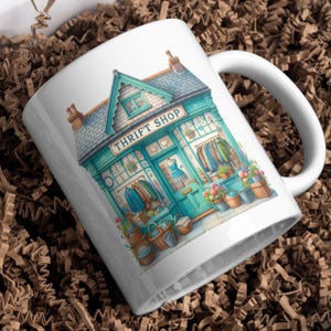 May include: White ceramic mug with a watercolour illustration of a teal Thrift Shop. The shop has a sign, clothing racks, and flower baskets. The mug has a white handle.