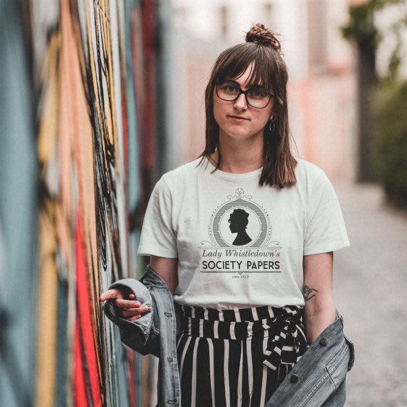 Pode incluir: Uma camiseta branca com um design gr&aacute;fico em preto e branco que apresenta uma silhueta de uma mulher e o texto "Lady Whistledown's Society Papers".
