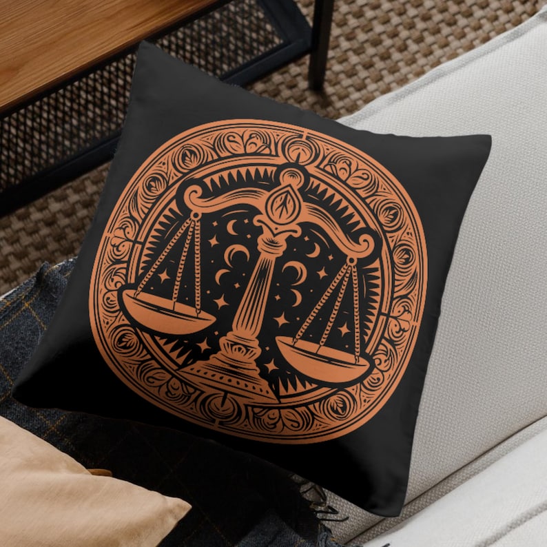 May include: A black throw pillow with an orange design of a scale with stars and crescent moons. The scale is surrounded by a decorative border.
