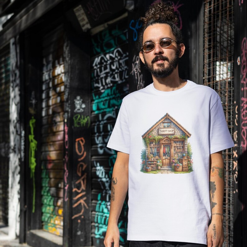 May include: White t-shirt with a colourful illustration of a thrift shop. The artwork shows a wooden building with a sign saying "THRIFT SHOP". The shop is surrounded by plants and various items, creating a charming and inviting scene.