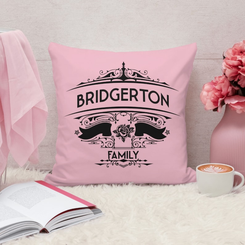 Pode incluir: Travesseiro rosa com um design preto que diz "Bridgerton Family" com uma rosa e trabalhos de rolagem ornamentados.