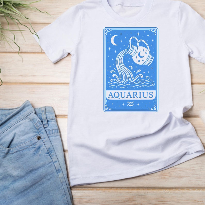 May include: White t-shirt with a blue graphic of the zodiac sign Aquarius. The graphic features a stylized image of a water pitcher pouring water into a body of water with stars and a crescent moon in the background. The text "AQUARIUS" is written below the image.