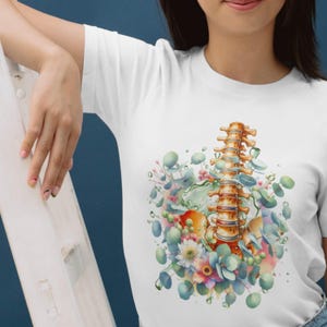 May include: White t-shirt featuring a watercolor illustration of a spine with floral and eucalyptus accents. The design includes a detailed depiction of the vertebrae, surrounded by soft pastel colors and green foliage.