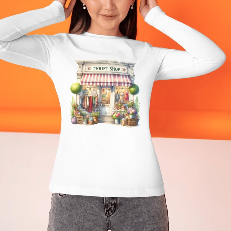 May include: White long-sleeved top with a watercolour illustration of a thrift shop. The shop has a red and white striped awning, with the words "THRIFT SHOP" above the entrance. The shop is surrounded by flowers and plants.