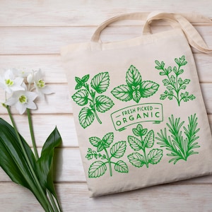 Może przedstawiać: Beżowa pł&oacute;cienna torba z zielonymi botanicznymi ilustracjami zi&oacute;ł i napisem "FRESH PICKED ORGANIC". Torba ma dwa uchwyty i jest umieszczona na jasnej drewnianej powierzchni. Białe kwiaty i zielone liście w tle.