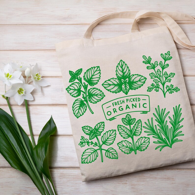 Może przedstawiać: Beżowa pł&oacute;cienna torba z zielonymi botanicznymi ilustracjami zi&oacute;ł i napisem "FRESH PICKED ORGANIC". Torba ma dwa uchwyty i jest umieszczona na jasnej drewnianej powierzchni. Białe kwiaty i zielone liście w tle.