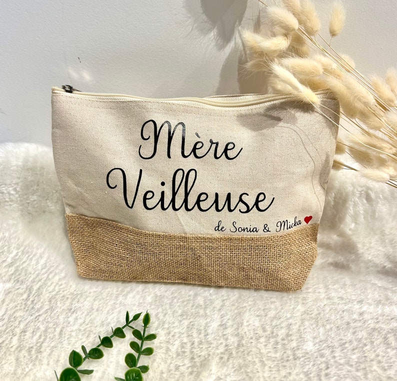 Op de afbeelding: Een beige canvas toilettas met een bruine jute bodem. De tas heeft zwarte tekst die "M&egrave;re Veilleuse de Sonia & Micka" luidt met een rood hart.