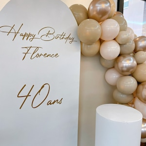 Op de afbeelding: Witte boog achtergrond met gouden belettering die "Happy Birthday Florence 40 ans" zegt en een ballonboog gemaakt van beige, crème en gouden ballonnen.