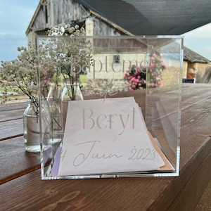 Könnte beinhalten: Eine transparente Acrylbox mit einer weißen Karte im Inneren. Die Karte trägt den Text "Bapteme de Beryl 4 Juin 2023". Die Box steht auf einem Holztisch mit weißen Blumen in Glasvasen.