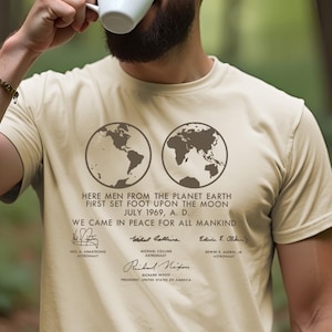 May include: A beige t-shirt with a graphic of two globes and the text "Here men from the planet Earth first set foot upon the Moon July 1969, A.D. We came in peace for all mankind."  The graphic also includes the signatures of Neil A. Armstrong, Michael Collins, and Edwin E. Aldrin Jr.