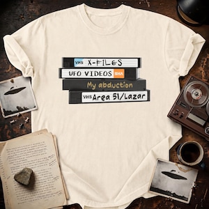 May include: A cream-colored t-shirt featuring a graphic of four stacked VHS tapes. The labels read X-FILES, UFO VIDEOS, My abduction, and Area 51/Lazar. The design is centered on the chest of the shirt, showcasing a retro aesthetic with text.