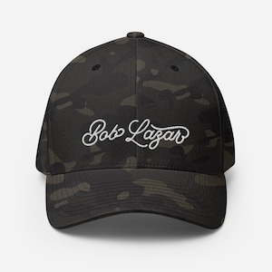 May include: A black and green camouflage baseball cap with white embroidered text that reads "Bob Lazar".