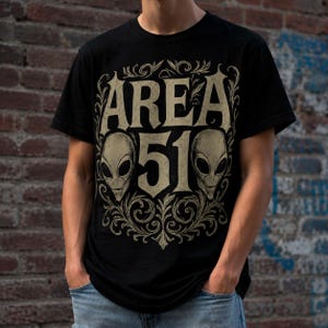 May include: A black short-sleeved t-shirt featuring a beige graphic print on the chest. The design displays the text AREA 51 in a stylised font, flanked by two alien heads, all surrounded by intricate, decorative scrollwork and filigree patterns.