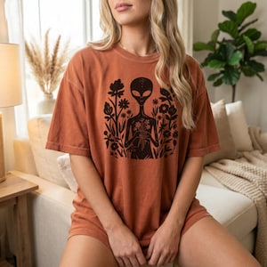 May include: Burnt orange t-shirt featuring a black graphic of an alien holding flowers, surrounded by floral elements. The shirt has short sleeves and a relaxed fit, perfect for casual wear. The design is centered on the front of the shirt.