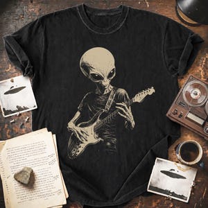 May include: A black short-sleeve t-shirt featuring a graphic print of an alien playing an electric guitar. The alien has a large head and dark eyes, and the guitar is detailed with strings and a neck. The shirt has a crew neckline and a relaxed fit.
