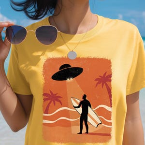 May include: Yellow t-shirt featuring a retro-style graphic of a UFO beaming down on a surfer holding a surfboard on a beach with palm trees. The shirt is made of a soft material.