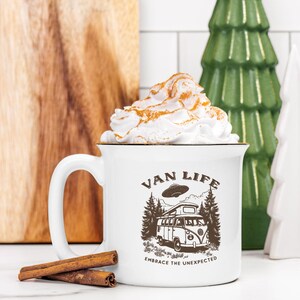 May include: White ceramic mug with a brown graphic of a van with a UFO above it. The text "Van Life" is above the van and "Embrace the Unexpected" is below. The mug has a white rim and a gold band around the top. The mug is filled with whipped cream.