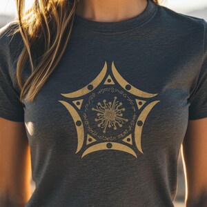 May include: Dark gray t-shirt featuring a gold-colored, stylized star design. The star has a central circular element with radiating lines and symbols around the perimeter. The shirt is made of a soft material.