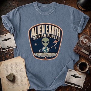 May include: A dusty blue t-shirt featuring a graphic emblem that reads ALIEN EARTH TOURISM BUREAU, APPROVED FOR VISITATION, SOL SYSTEM, SECTOR 03. The design includes a green alien head, a depiction of Earth, and a small flying saucer icon.