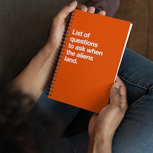 May include: An orange spiral-bound notebook with the text "List of questions to ask when the aliens land." The notebook is held in a person's hands, with the person wearing blue jeans.