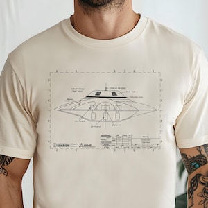 May include: Cream-colored t-shirt featuring a detailed blueprint of a UFO. The design includes technical labels and a grid, with the word "ENERGY" and other text at the bottom. A unique graphic tee.