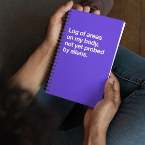 May include: A purple spiral-bound notebook held in a person's hands. The notebook's cover features the text: "Log of areas on my body, not yet probed by aliens." The notebook is open, revealing white pages.