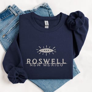 May include: Navy blue sweatshirt with the text "ROSWELL NEW MEXICO" in silver, along with a graphic of a flying saucer. The sweatshirt is folded on top of a pair of blue jeans.
