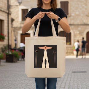 May include: Beige tote bag featuring a retro-style graphic of a UFO beaming down a person. The design includes a black background, a light beam, and circular accents. The bag has long handles.