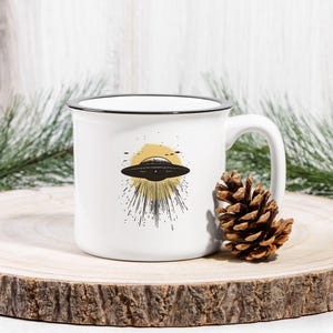 May include: White ceramic mug with a black rim and handle. The mug features a black and white illustration of a UFO with a yellow sun behind it.