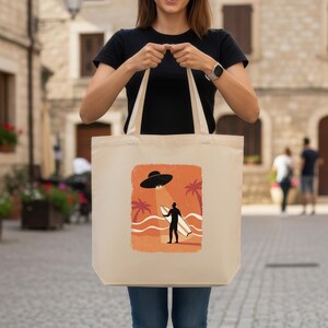 May include: Beige tote bag featuring a graphic of a UFO beaming down on a surfer holding a surfboard on a beach with palm trees. The bag is held by a person wearing a black shirt and blue jeans.