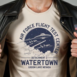 May include: A beige t-shirt with a blue and white graphic design featuring a UFO, two fighter jets, and the text "Air Force Flight Test Center, Detachment 3, Watertown, Groom Lake Nevada".