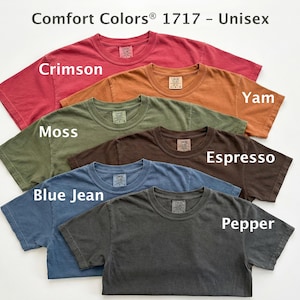 May include: A collection of Comfort Colors 1717 unisex t-shirts in various colors, including Crimson, Yam, Moss, Espresso, Blue Jean, and Pepper. Each shirt has a crew neck and short sleeves, with the color names displayed on the shirts.