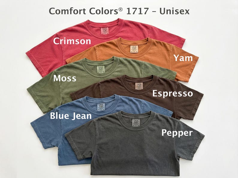 May include: A collection of Comfort Colors 1717 unisex t-shirts in various colors, including Crimson, Yam, Moss, Espresso, Blue Jean, and Pepper. Each shirt has a crew neck and short sleeves, with the color names displayed on the shirts.