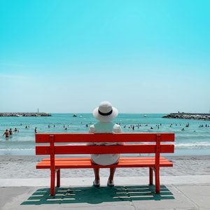 May include: A person wearing a white hat and dress sits on a bright orange bench, facing the ocean. The bench is on a concrete walkway, with a sandy beach and blue water in the background. People are swimming in the sea under a clear sky.