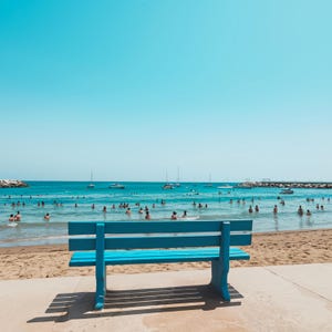 May include: A bright blue wooden bench sits on a concrete surface, casting a shadow on a sunny beach. The ocean is filled with people swimming, with boats in the distance. The sky is a clear, vibrant blue.