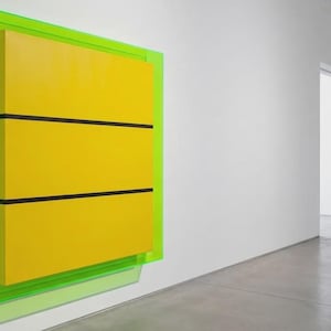 May include: A modern art piece featuring a bright yellow rectangular form with three horizontal black stripes, encased in a neon green acrylic frame. The artwork is mounted on a white wall in a minimalist gallery setting.