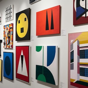 May include: A gallery wall displays a collection of framed abstract paintings. The artworks feature geometric shapes, bold colors like red, yellow, and blue, and various compositions. The paintings vary in size and style, creating a diverse art exhibition.