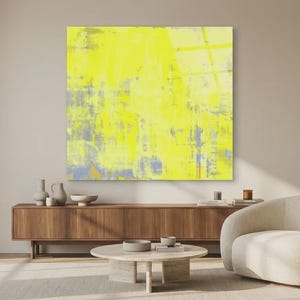May include: A large, square abstract painting dominates the scene, featuring a vibrant yellow color palette with hints of blue and gray. The artwork hangs above a wooden cabinet, complemented by a cream-colored armchair and a round coffee table. Decorative vases and bowls add to the room's aesthetic.