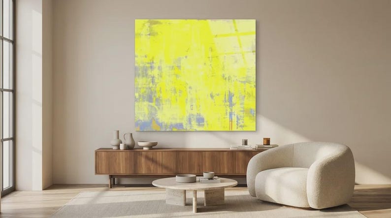 May include: A large, square abstract painting dominates the scene, featuring a vibrant yellow color palette with hints of blue and gray. The artwork hangs above a wooden cabinet, complemented by a cream-colored armchair and a round coffee table. Decorative vases and bowls add to the room's aesthetic.