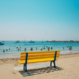 May include: A yellow bench with blue legs sits on a sandy beach, overlooking a calm, turquoise ocean. Several people are swimming in the water, and boats are visible in the distance under a clear, blue sky.
