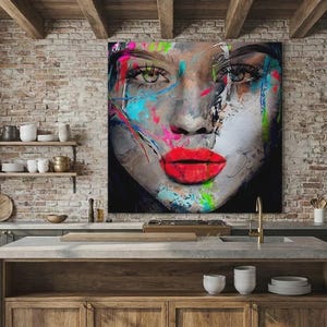 May include: A large canvas art piece featuring a woman's face with vibrant, abstract paint strokes in shades of red, blue, green, and white. The artwork has bold red lips and expressive eyes, creating a striking visual impact. The art is displayed in a kitchen setting.