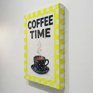 Könnte beinhalten: Ein rechteckiges Wandkunstwerk mit den Worten "COFFEE TIME" in schwarzen Buchstaben. Unter dem Text befindet sich eine Tasse Kaffee auf einer Untertasse. Das Kunstwerk hat einen gelb-weiß karierten Rand.