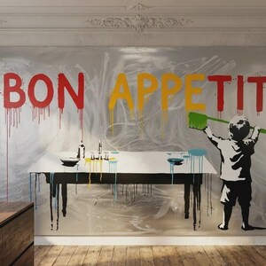 May include: A large wall art piece in a kitchen setting. The artwork features the words "BON APPETIT" in red, yellow, and red, with paint drips. A child figure is depicted holding a green brush. A table with dishes and bottles is in front of the artwork.