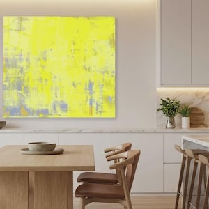 May include: A modern kitchen with a large abstract yellow painting. The painting has a textured appearance with hints of gray and blue. The kitchen features a marble countertop, wooden chairs, and pendant lights.