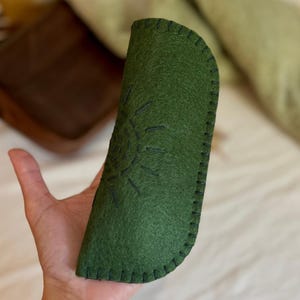May include: A green felt glasses case with a stitched sunburst design. The case is hand-stitched with black thread and has a curved top. The brand name "folka concept" is written in cursive on the bottom right.