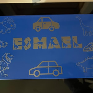 Puede incluir: Un objeto rectangular azul con ilustraciones doradas de dinosaurios y coches. El nombre "ESMAEL" se muestra en letras doradas con siluetas de dinosaurios. El diseño incluye varios contornos de dinosaurios y coches.